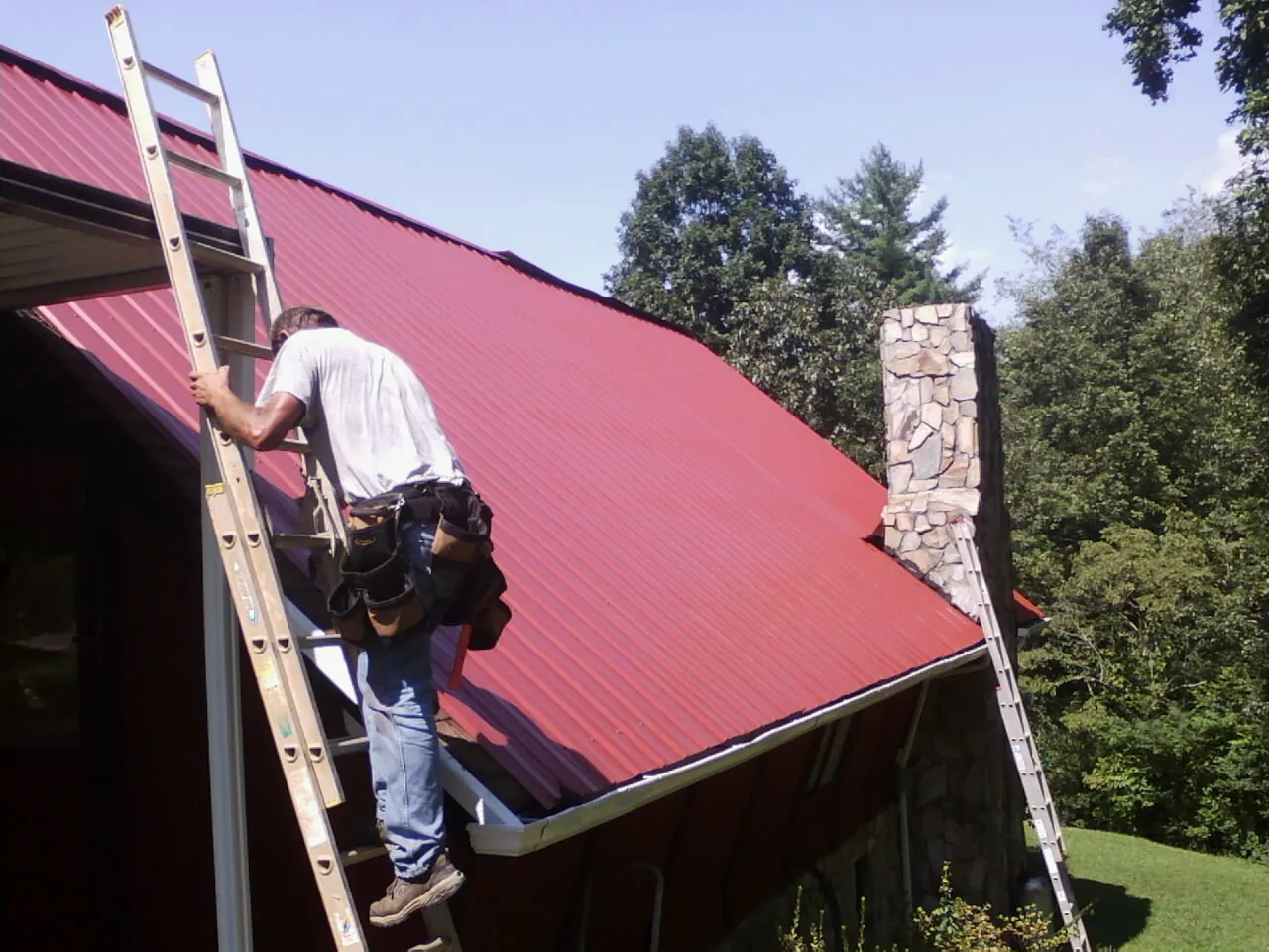 Roofer on a ladder beside a red metal roof and stone chimney — Soffit & Fascia Repair by Roofing Brothers in Cedaredge